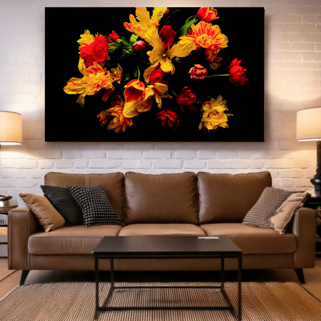 Een levendige foto van rode en gele bloemen tegen een zwarte achtergrond, getoond als kunstwerk boven een bruine leren bank in een woonkamer. De bank heeft diverse sierkussens in zwart-wit patronen en neutrale tinten. Voor de bank staat een donkere salontafel op een gestreept tapijt. De witte bakstenen muur en twee wandlampen aan weerszijden van de foto maken het interieur compleet.