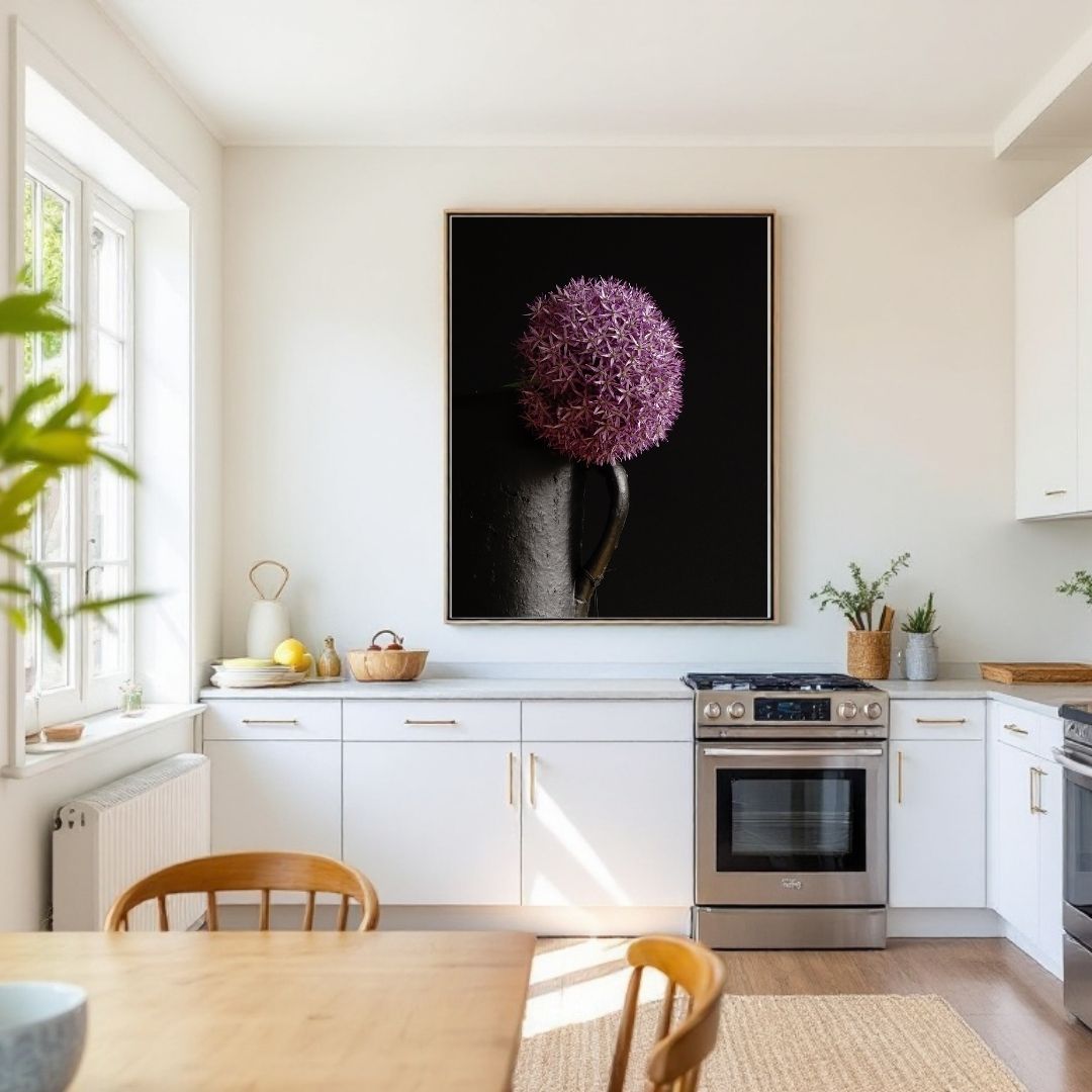 Lichte, moderne keuken met witte kasten en werkblad. Een groot ingelijst kunstwerk van een paarse allium bloem tegen een zwarte achtergrond hangt boven het fornuis. Houten eettafel met stoelen op de voorgrond, grote ramen links die daglicht binnenlaten en wat groen zichtbaar aan de zijkanten voor een natuurlijke toets.