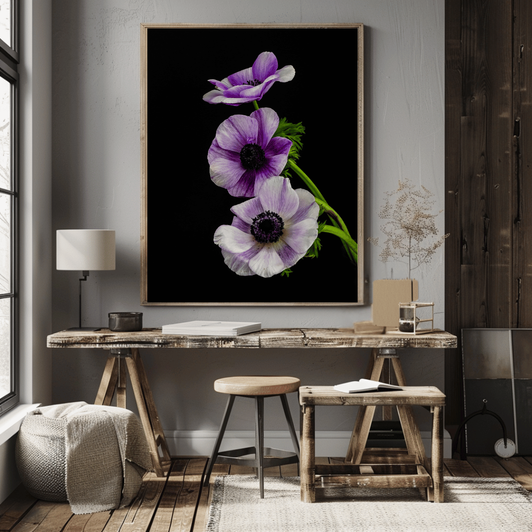 Een grote ingelijste poster van paarse anemoonbloemen op een zwarte achtergrond. De poster hangt boven een rustieke houten bureau of tafel in een kamer met een minimalistische, natuurlijke inrichting. De ruimte heeft houten vloeren, een grijze muur, en bevat enkele decoratieve elementen zoals een kleine tafellamp, gedroogde planten, een geweven poef en een klein houten krukje. Het geheel straalt een rustige, elegante sfeer uit met natuurlijke materialen en een opvallend bloemenkunstwerk als focuspunt