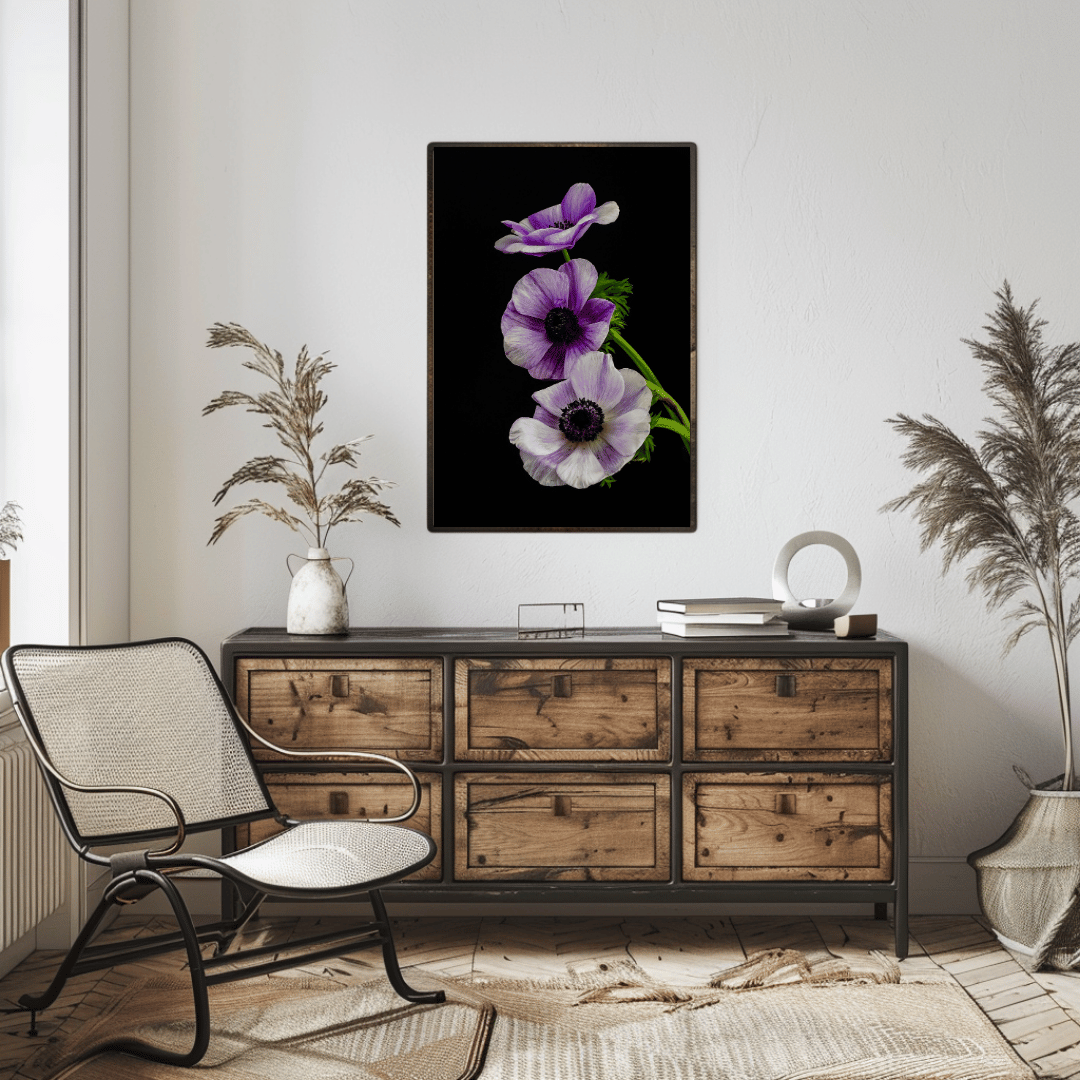 Een ingelijste poster met drie paarse anemoonbloemen op zwarte achtergrond hangt aan een witte muur boven een rustieke houten dressoir met zes lades. Links staat een elegante zwarte stoel met wit geweven zitting en leuning. Aan weerszijden van het dressoir staan pampasgraspluimen in vazen als decoratie. Op het dressoir liggen enkele boeken en een ronde decoratieve sculptuur. De ruimte heeft een lichte houten vloer met een natuurlijk geweven tapijt