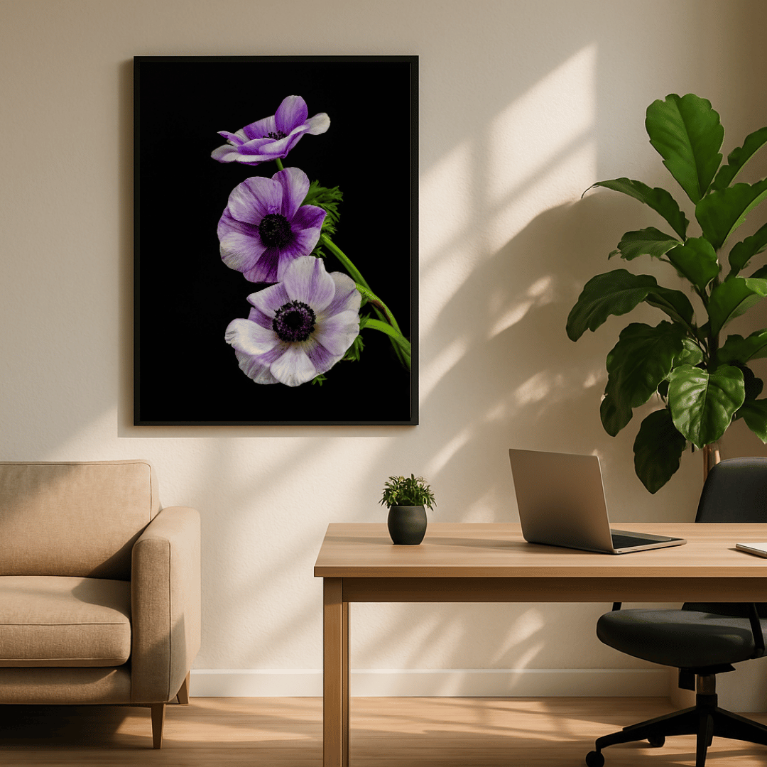Een moderne werkruimte of thuiskantoor met een ingelijste kunstafbeelding aan de muur waarop drie paarse anemoonbloemen met donkere harten tegen een zwarte achtergrond te zien zijn. De bloemen zijn verticaal gerangschikt op een groene stengel. In de ruimte staat een houten bureau met een grijze laptop en een kleine groene plant in een donkere pot. Bij het bureau staat een zwarte bureaustoel op wieltjes. Aan de linkerkant van het beeld is een deel van een beige bank of fauteuil zichtbaar. Rechts in de hoek s